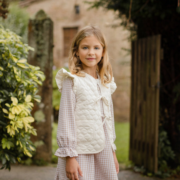 Young girl in a checkered dress standing outdoors with greenery and a building in the background