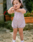 A baby girl wearing a short pink romper with a floral pattern, including delicate straps with bows, and a matching hat.