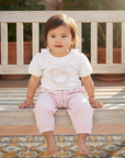 Asian baby girl in pink pants and white teddy bear top on wooden bench over Spanish tiles