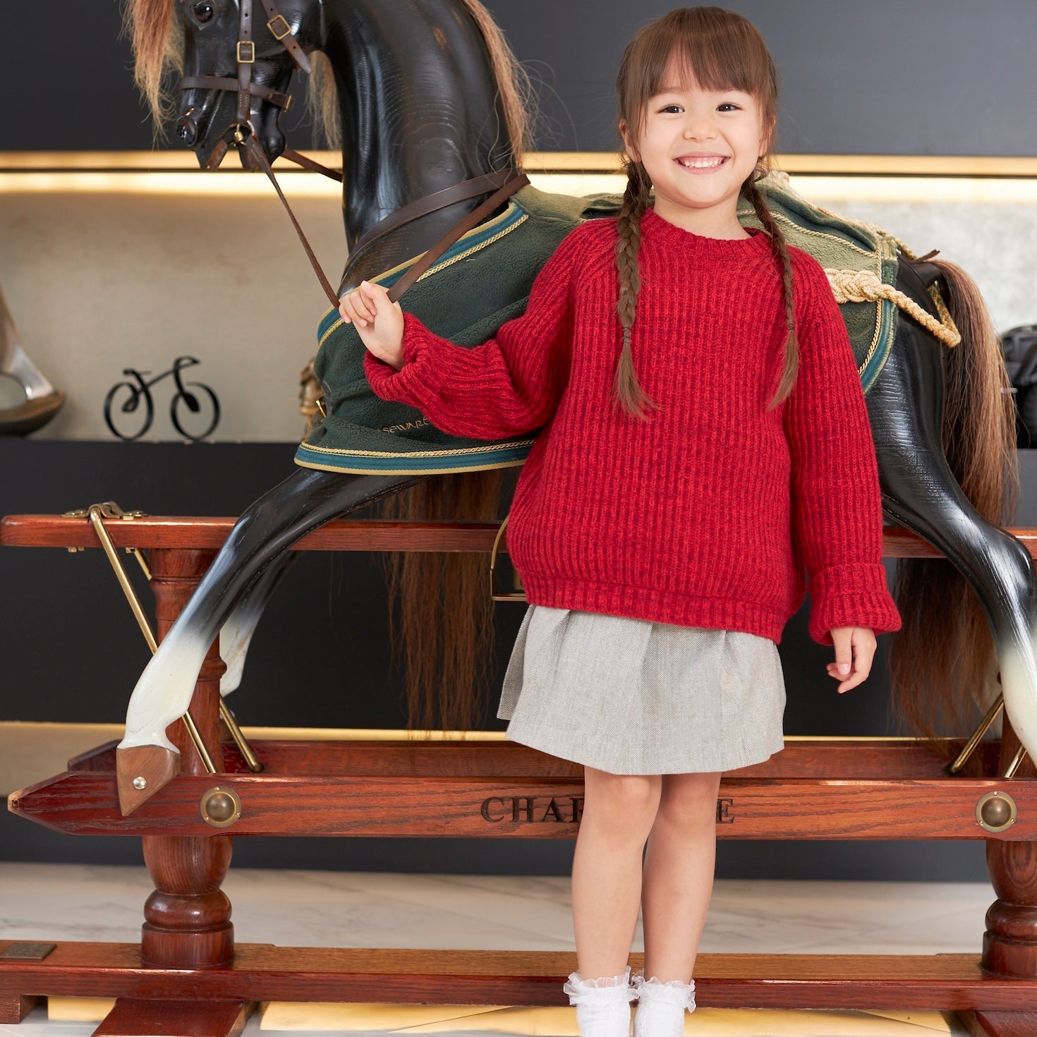 Child in a red sweater standing next to a decorative horse statue indoors.