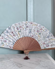 Decorative hand fan with floral pattern on a light gray surface against a light blue wall.
