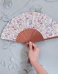 Hand holding a floral-patterned fan against a decorative wall.