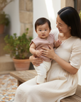 Asian baby girl in dusty pink knitted vest and white pants held by Asian mother on wooden bench over Spanish tiles
