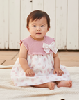 Asian baby girl in pink and white floral dress seated outdoors with white wooden background